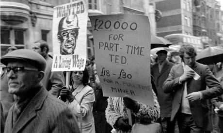 Black and white photo of a protest march, featuring individuals holding signs advocating for a living wage, one sign reads '£20,000 for part-time Ted, £18-£30 for a full-time miner.' People wearing coats and hats, with umbrellas in the background.